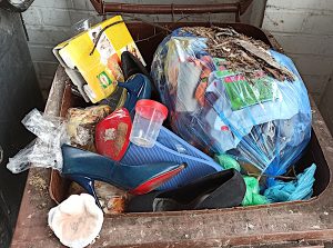 Na zdjęciu przedstawiono brązowy pojemnik na odpady ulegające biodegradacji w którym znajdują się odpady innych typów: plastik, papier lakierowany, buty itp.