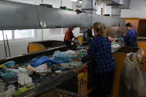 Na zdjęciu przedstawiono ruchomą taśmę z odpadami. Pracownicy stojący przy taśmie wybierają poszczególne frakcje odpadów.