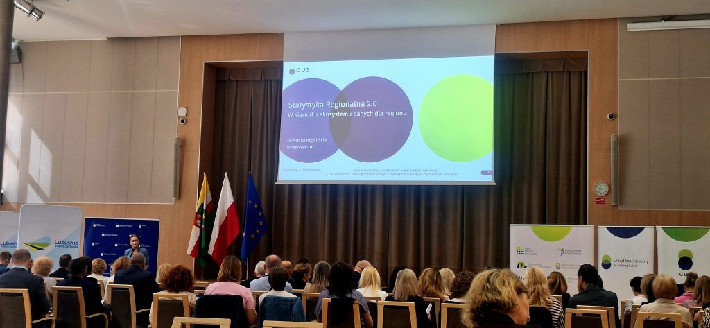 Sala konferencyjna z uczestnikami spotkania widziana od tyłu. Na środku ekran z tytułem prezentacji „Statystyka Regionalna 2.0 w kierunku ekosystemu danych dla regionu”. Po lewej stronie flagi: województwa lubuskiego z herbem, Polski oraz Unii Europejskiej. Po prawej stronie rollup’y Urzędu Statystycznego.